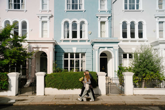 A stylish couple walks past colourful pastel townhouses on a sunny London street, reflecting the sophisticated, urban lifestyle behind Savyll non-alcoholic cocktails.